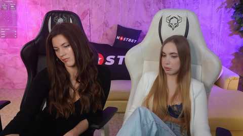 Media: Video of two women in gaming chairs; one with long wavy hair, black top, and blue jeans; another with long blonde hair, white top, and blue jeans, both looking bored.