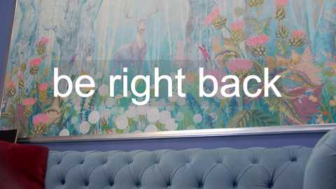 Media: Video of a vibrant, floral-patterned canvas painting with a blue velvet tufted sofa below. Text reads \"be right back.\" The room's walls are painted a soft lavender.