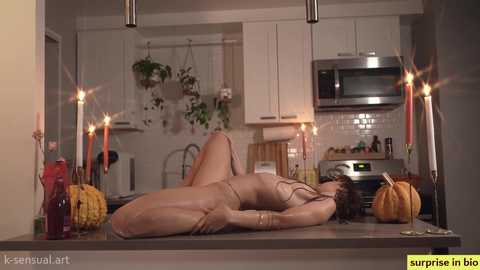 Media: A video of a naked woman lying on a kitchen counter, surrounded by candles, pumpkins, and kitchen appliances, with a serene, intimate ambiance.