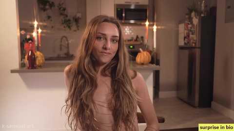 Media: A video of a young woman with long, wavy brown hair, sitting topless in a modern kitchen. Pumpkins and decorative items on the counters.