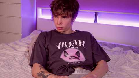 Media: Video of a young man with short, dark hair, wearing a black Nirvana t-shirt, lying on a bed with white sheets, in a dimly lit bedroom with purple lighting.