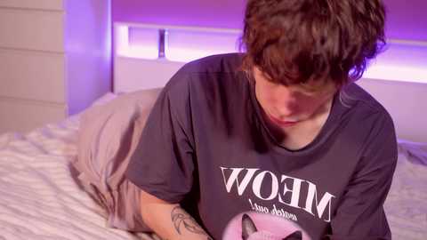 Media: Video of a young woman with short brown hair lying on a bed, wearing a dark T-shirt with white text, looking at a pink cat toy.