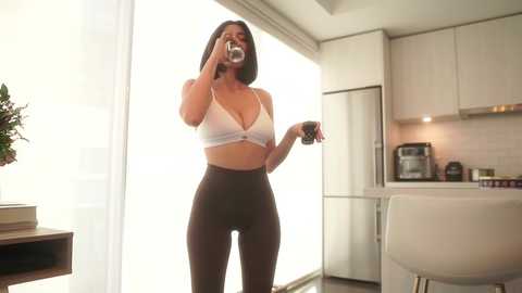 Media: Video of a fit woman with medium skin tone and long dark hair, wearing a white sports bra and black leggings, drinking coffee in a modern, light-filled kitchen with stainless steel appliances.