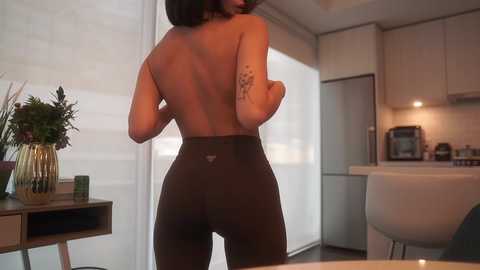 Media: Video of a topless woman in brown leggings, standing in a modern kitchen with a tattoo on her left arm.