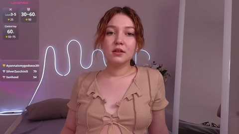 Media: Video of a young woman with fair skin, light brown hair, wearing a beige top, standing in a dimly lit room with a neon \"U\" sign and social media stats displayed.