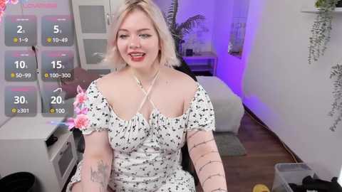 Media: A video of a smiling, blonde woman with tattoos, wearing a white floral dress, sitting on a couch in a modern, dimly lit living room with purple accents.