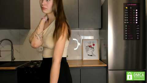 Media: Video of a young, slim, light-skinned woman with long brown hair, wearing a beige sleeveless top and black skirt, in a modern kitchen with stainless steel appliances and marble backsplash.