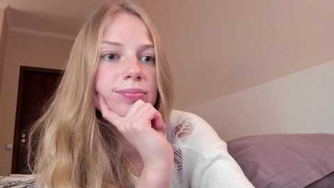 Media: A video of a young Caucasian woman with long, blonde hair, resting her chin on her hand, wearing a white lace top, in a beige room with a brown door in the background.