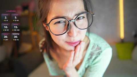 Media: Video of a young woman with glasses, fair skin, and brown hair, resting her chin on her hand, in a modern, dimly lit room.