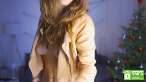 Media: A video of a woman with long, wavy brown hair, wearing a tan jacket, standing in a dimly lit room with a Christmas tree adorned with red and silver ornaments in the background.