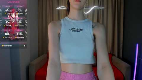 Video of a slender, fair-skinned woman with auburn hair, wearing a light blue crop top and pink shorts, standing indoors in front of a virtual reality setup.