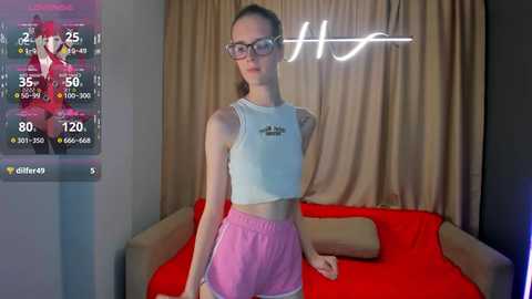 Video of a slender, fair-skinned young woman with glasses, wearing a white crop top and pink shorts, standing in a modern, dimly lit bedroom with beige curtains and a red bed.