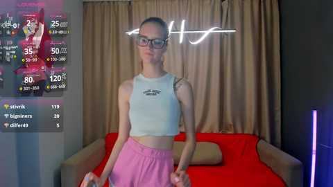 Video of a slender, fair-skinned woman with glasses and pink shorts, standing in a modern bedroom with beige curtains and a red bed.