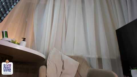 Media: Video of a cozy living room with beige curtains, a white cushion, a small green plant, and a round wooden table with a green bottle.