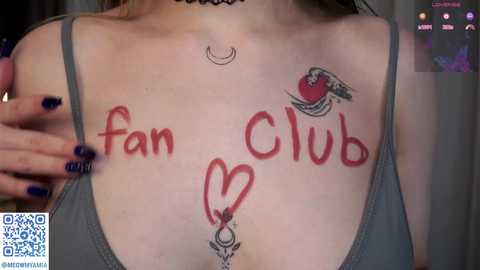 Media: Video of a fair-skinned woman with a choker and tattoos, including a heart and \"fan club\" written in red ink on her chest. Her nails are painted dark blue.