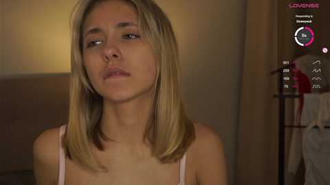 Media: Video of a young woman with light skin and straight, shoulder-length blonde hair, wearing a pink tank top. She has a neutral expression. Background includes a beige wall and a partially visible bed.