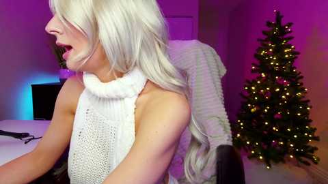 Media: Video of a blonde woman in a white knitted sweater, mouth open, in a dimly lit room with a lit Christmas tree and purple lighting.