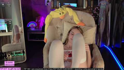 Media: A video of a young woman in a plush armchair, wearing white thigh-high socks, surrounded by plush toys and colorful lights in a dimly lit room.