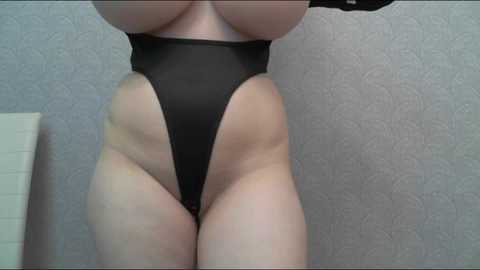 Media: A video of a fair-skinned woman with a curvy physique wearing a high-cut black thong bodysuit, highlighting her ample buttocks and toned thighs against a textured, light gray wall.