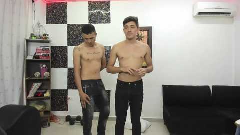 Media: Video of two shirtless, lean Asian men in tight black jeans, standing in a modern living room with black-and-white checkered wall, bookshelf, and black couch.