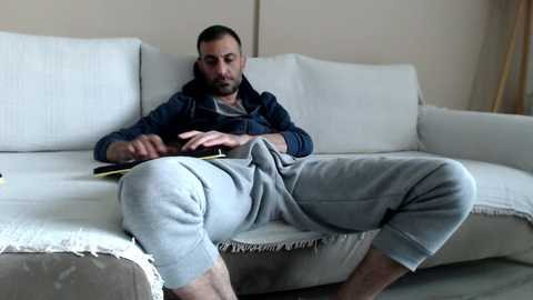 Media: Video of a bearded man with short dark hair, wearing a blue hoodie and gray sweatpants, sitting on a white couch, reading a book, in a minimalist living room.