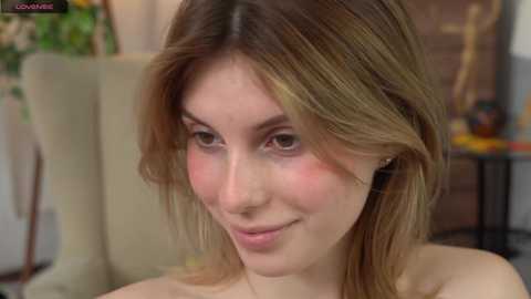 Media: Video of a young Caucasian woman with light skin and shoulder-length, wavy brown hair, smiling softly. She wears a strapless top, with a blurred background featuring colorful artwork and furniture.