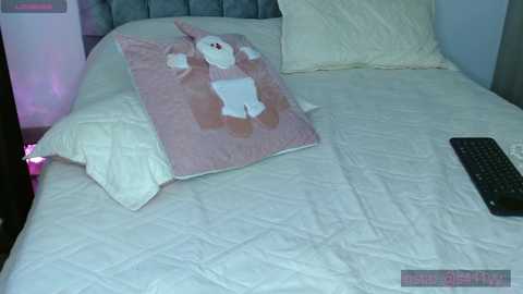 Media: Video of a neatly made bed with a plush rabbit pillow and a black keyboard on the white quilted comforter. The backdrop features a light blue wall and a bedside lamp with a pink glow.