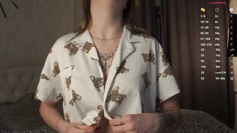 Media: Video of a light-skinned woman with a tattoo on her arm, wearing a white, short-sleeved, floral-patterned shirt, partially unbuttoned, revealing a lace bra, in a dimly lit bedroom.