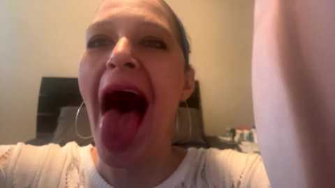 Media: A video of a young woman with fair skin, blue eyes, and dark hair tied back. She has a wide-open mouth with her tongue sticking out, wearing a white top. The background shows a beige wall and indistinct furniture.