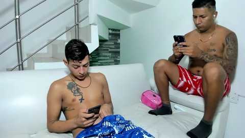 Media: Video of two shirtless Latino men with tattoos; one in red shorts, the other in blue, sitting on a white couch. Both are engrossed in their smartphones.