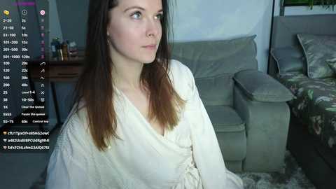 Media: Video of a Caucasian woman with long brown hair, wearing a white robe, sitting in a living room with a gray couch and floral pillows, with a computer screen displaying social media notifications in the background.