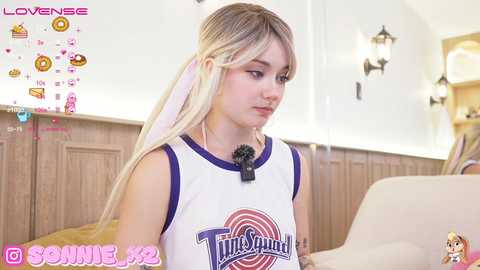 Media: Video of a young woman with long blonde hair, wearing a \"Looney Tunes\" basketball jersey, sitting indoors. The background features wooden paneling and a lamp. Text overlays indicate \"Lovense\" and \"Sunny_69.\