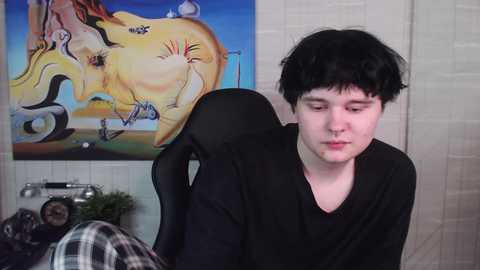 Media: Video of a young, pale-skinned man with short black hair, wearing a black shirt, sitting in a black chair. Background features a colorful abstract painting and a plaid blanket.