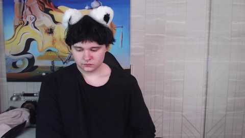 Media: A video of a young person with short black hair wearing a panda-themed headband and black hoodie, sitting in a room with a colorful abstract painting and beige tiled walls.