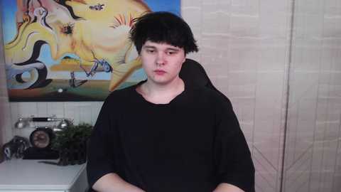 Media: Video of a young man with messy black hair in a black t-shirt, seated in a barber chair, with a colorful, abstract painting of a woman and a horse on a beige wall.