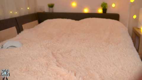 Media: A video of a cozy bedroom with a large, fluffy white blanket covering a bed, surrounded by warm, glowing fairy lights on a white wall. The room is dimly lit, giving a soft, inviting atmosphere.