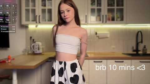 Media: Video of a young girl with long brown hair in a white crop top and black heart-patterned skirt, standing in a modern kitchen with white cabinets and a wooden countertop.