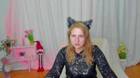 Media: Video of a fair-skinned, blonde woman with cat ears, wearing a black lace dress, sitting on a white couch. Background includes a pink light, a red Santa hat, a white shelf with flowers, and a vase.