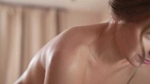 Media: Video of a topless woman with fair skin, brown hair tied back, her profile showing smooth skin and a slender neck. Background features beige curtains.