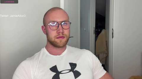 Media: Video of a bald, bearded, muscular man with glasses, wearing a white Under Armour shirt, standing in a doorway with a headset on, in a simple, neutral-toned room.