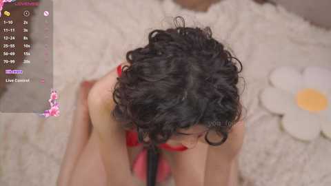 Media: Video of a woman with curly black hair and fair skin, wearing a red bra, kneeling on a fluffy white rug with a white flower beside her.