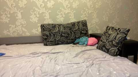 Media: Video of a neatly made bed with light gray sheets and two decorative pillows featuring intricate floral patterns in gold and black. Background shows a wallpaper with similar floral designs in muted tones.