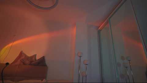 Media: A video of a minimalist, dimly lit bedroom with a beige sofa, decorative pillows, and two modern white floor lamps. A vibrant orange and pink light casts a warm glow on the walls, creating a serene ambiance.