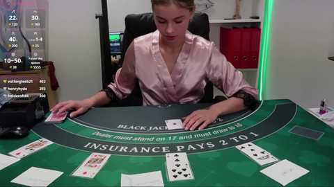 Media: A video of a young woman, with fair skin and blonde hair tied back, in a pink satin robe, seated at a green blackjack table with cards and chips, in a dimly lit, modern room with a TV screen showing game stats.