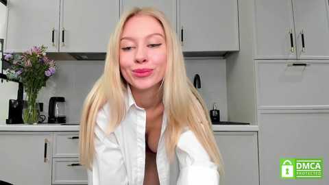 Media: Video of a blonde woman with fair skin and pink lips, wearing a white blouse, in a modern kitchen with white cabinets and purple flowers.