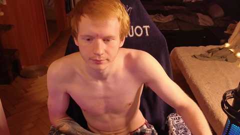 Media: Video of a young, shirtless, fair-skinned man with short red hair, sitting on a bed in a dimly lit room, wearing patterned shorts.