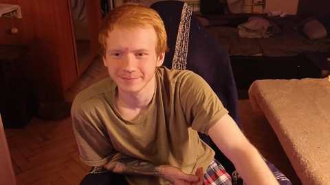 Media: Video of a young man with red hair and fair skin, wearing a green T-shirt and plaid pajama bottoms, sitting on a bed in a dimly lit room with scattered clothes and a wooden floor.