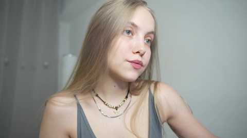 Media: Video of a young Caucasian woman with straight blonde hair, wearing a gray spaghetti-strap top, layered gold necklaces, and subtle makeup. She gazes slightly to the left, against a plain, light gray background.