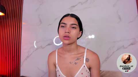 Media: Video of a young woman with medium skin tone, black hair tied back, wearing a white spaghetti-strap tank top. She has small breasts, visible tattoos on her chest, and a circular ring light in the background.