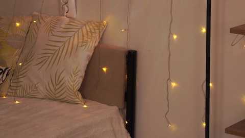 Media: Video of a cozy bedroom with a bed covered in a white blanket, a pillow with a golden leaf pattern, and warm, glowing fairy lights adorning the beige wall.